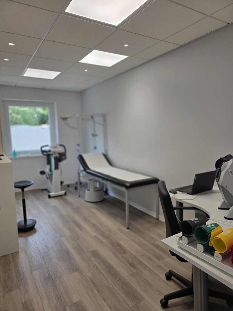 Der Raum ist ein Behandlungszimmer mit einem Untersuchungstisch, einem Bürostuhl und einem Tisch mit medizinischen Geräten. Es gibt auch eine Trainingsmaschine und farbige Therapiegeräte auf dem Tisch. Tageslicht fällt durch ein Fenster.
