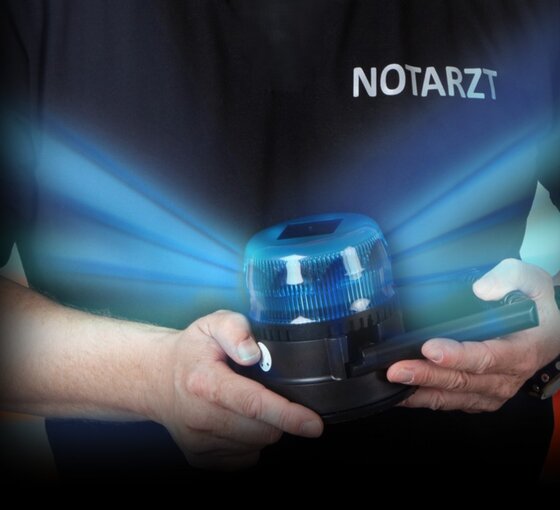 Ein Notarzt hält ein blaues Blitzlicht in den Händen. Das Licht strahlt nach außen und erzeugt einen dramatischen Effekt. Der Arzt trägt ein schwarzes Shirt mit dem Wort "NOTARZT". Der Hintergrund ist unscharf und verstärkt die Aufmerksamkeit auf das Blitzlicht.