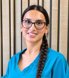 Eine lächelnde Person mit langen, geflochtenen Haaren trägt ein türkisfarbenes Oberteil und eine Brille. Sie steht vor einer Wand mit Holzlatten.