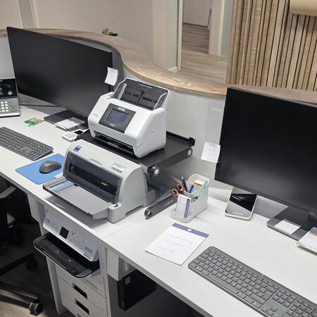 Ein modernes Büro mit zwei großen Monitoren, einem Drucker und einem Scanner. Auf einem Schreibtisch sind ein Telefon, Büromaterialien und Notizen platziert. Der Raum ist minimalistisch und gut organisiert, mit Holz- und neutralen Farbtönen.