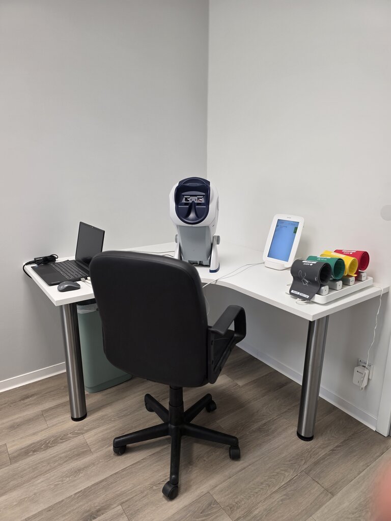 Ein moderner Arbeitsplatz mit einem Schreibtisch, einem ergonomischen Bürostuhl, einem Laptop links, einem medizinischen Gerät in der Mitte und einem Touchscreen-Rechner rechts. Farbcodierte Zubehörteile liegen auf dem Tisch. Der Raum hat einen hellen, minimalistischen Stil.