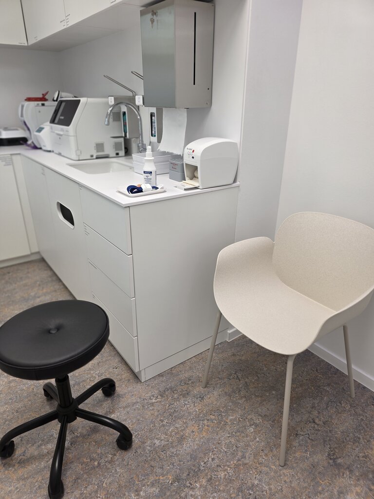 Eine helle Laborumgebung mit einem weißen Tisch, einer Küchenzeile und zwei Stühlen. Ein Drehstuhl mit schwarzem Polstersitz und ein weißer, moderner Stuhl sind sichtbar. Auf dem Tisch stehen ein Laborgerät und andere Utensilien. Der Boden ist grau-beige.