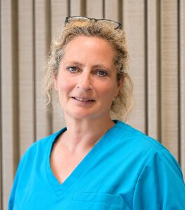 Eine Frau mit blonden Locken trägt ein türkisfarbenes Oberteil. Sie schaut freundlich in die Kamera und steht vor einer hellen, strukturierten Wand.