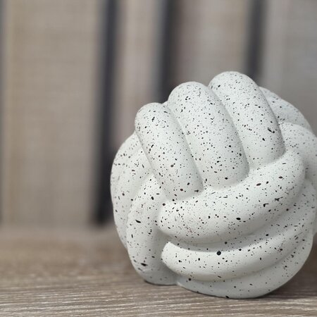 Eine weiße, knotenähnliche Skulptur mit schwarzen Sprenkeln, die auf einer Holzoberfläche liegt. Der Hintergrund ist unscharf und zeigt helle, vertikale Streifen.
