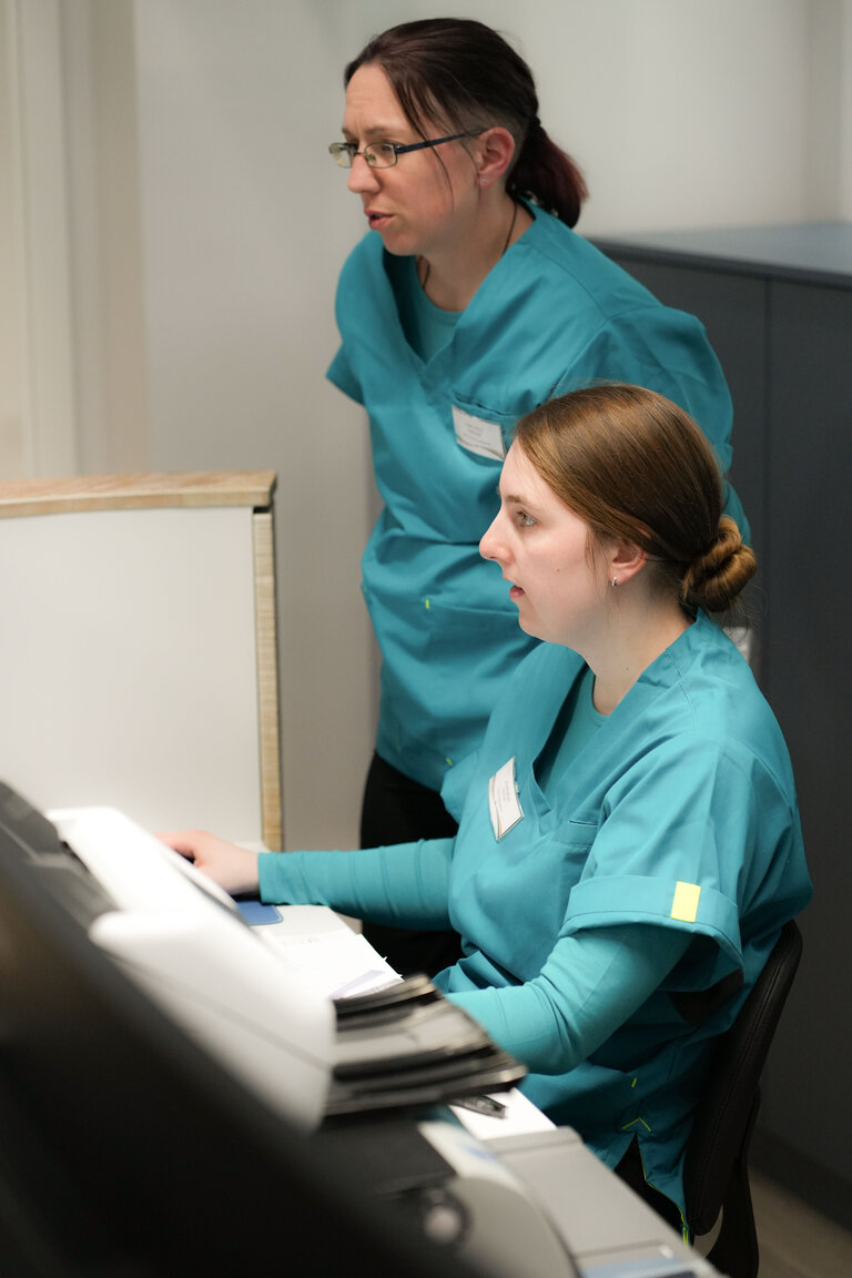 Zwei Frauen in blauen Arbeitskleidung stehen und sitzen an einem Schreibtisch. Eine Frau schaut auf einen Computerbildschirm, während die andere hinter ihr steht und aufmerksam zusieht. Der Raum scheint hell und modern zu sein.
