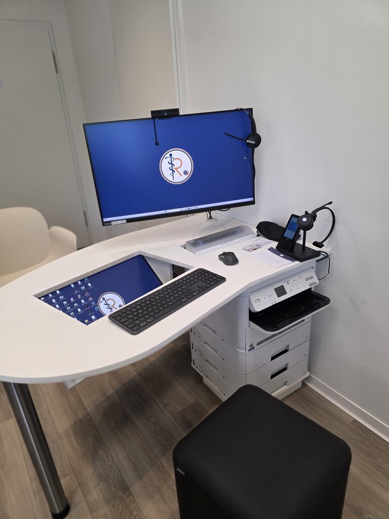 Ein moderner Schreibtisch mit einem großen Computerbildschirm, Tastatur und Maus. Rechts steht ein Drucker auf einem weißen Getränkeschrank, daneben ist ein Mikroskop. Der Raum wirkt hell und einladend.