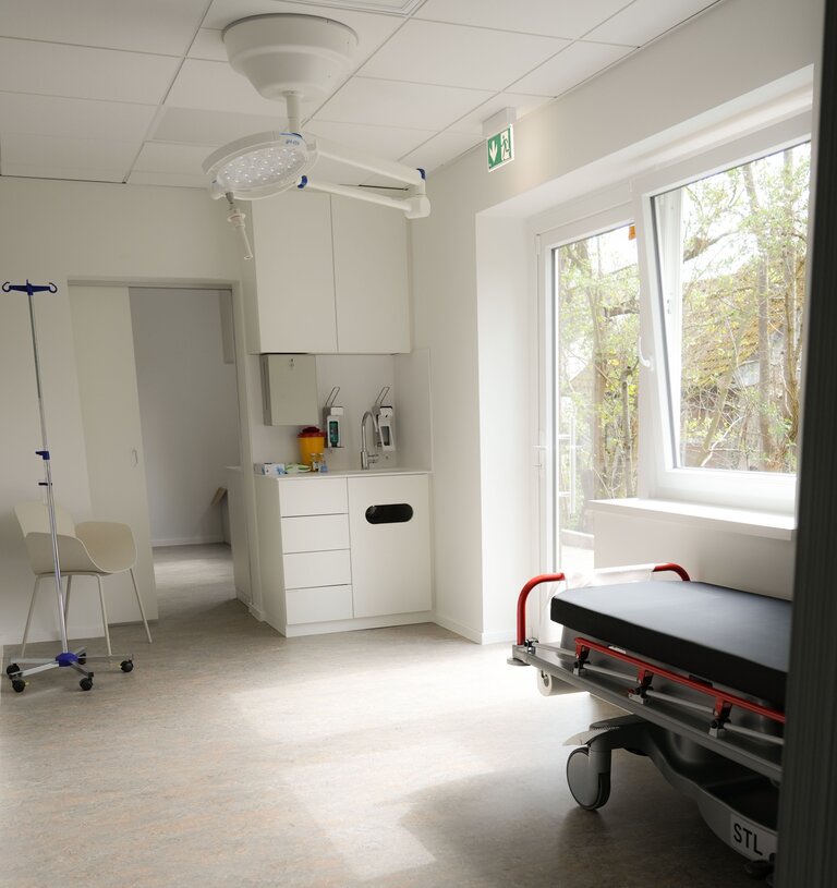 Der Innenraum eines modernen medizinischen Raums, ausgestattet mit einem Behandlungsbett, einem kleinen Tisch, einer medizinischen Geräteabdeckung und einem Fenster mit Blick auf die Außenanlage. Im Hintergrund sind medizinische Utensilien sowie ein Stuhl zu sehen.