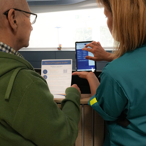 Ein älterer Mann mit Brille schaut auf ein Informationsblatt, während eine Frau in blauer Arbeitskleidung ein Smartphone hält und darauf tippt. Die Szene vermittelt einen interaktiven Austausch zwischen den beiden Personen.