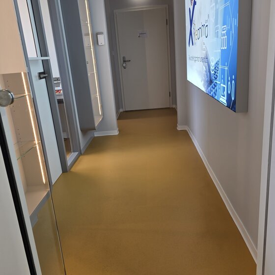 Ein Flur mit gelbem Boden, der zu einer Tür am Ende führt. An einer Wand befindet sich ein Bildschirm mit grafischen Darstellungen. Gegenüber ist ein Regal mit Glasfront, das leer erscheint. Das Licht ist hell und freundlich.