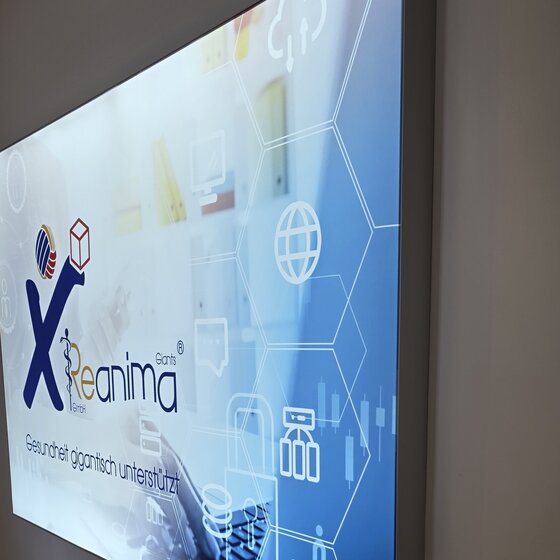 Ein Digitalbildschirm an einer Wand zeigt das Logo von "Xeanima GmbH" und den Slogan "Gesundheit ganzheitlich unterstützt" auf einem hellen, modernen Hintergrund mit verschiedenen grafischen Elementen.