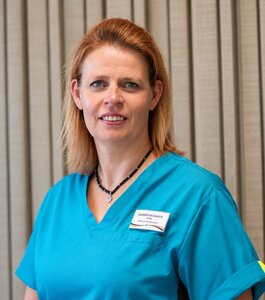 Eine Frau mit blonden Haaren trägt ein türkisfarbenes Oberteil und steht vor einer beigefarbenen Wand. Sie lächelt und hat eine Kette um den Hals. Auf ihrer Brust befindet sich ein Namensschild.