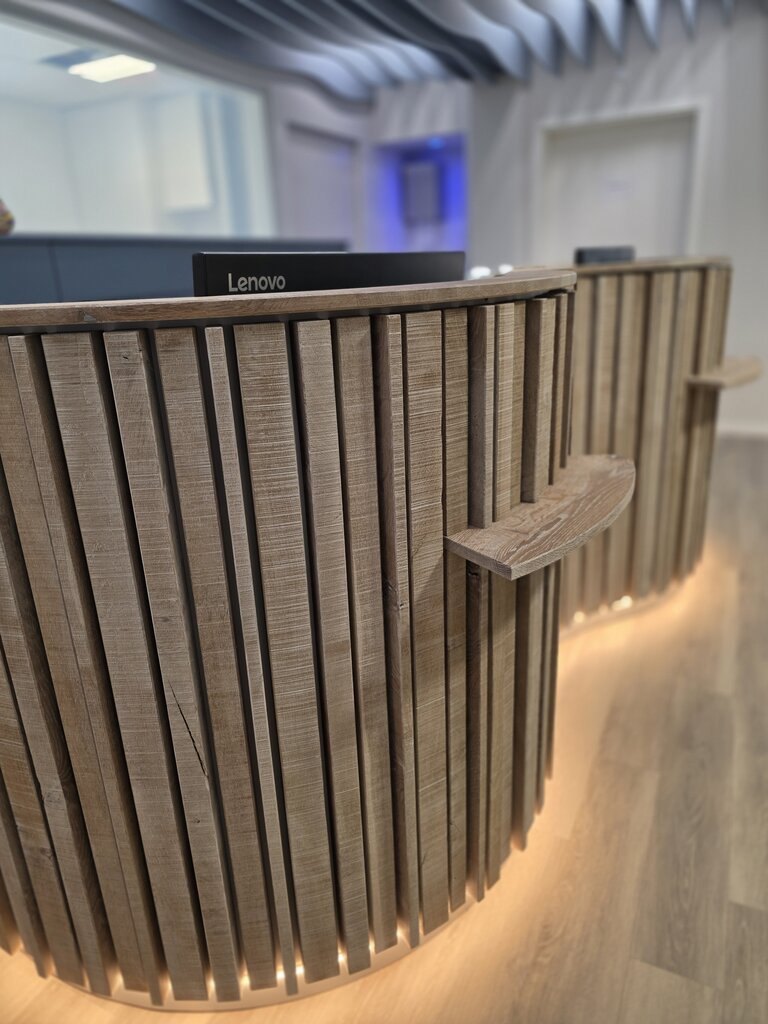 Ein moderner Empfangstresen mit Holzverkleidung und sanfter Beleuchtung. Der Tresen hat vertikale Holzlamellen und eine durchgehende Ablagefläche. Der Schriftzug "Lenovo" ist sichtbar. Der Raum wirkt hell und einladend.