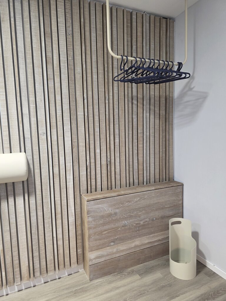 Ein minimalistischer Raum mit einer horizontalen Holzlattenwand. An der Wand hängt ein schwarzes Kleidergestell. Darunter steht ein Holzschrank mit zwei Schubladen und rechts daneben befindet sich ein minimalistischer, runder Behälter aus hellem Material.