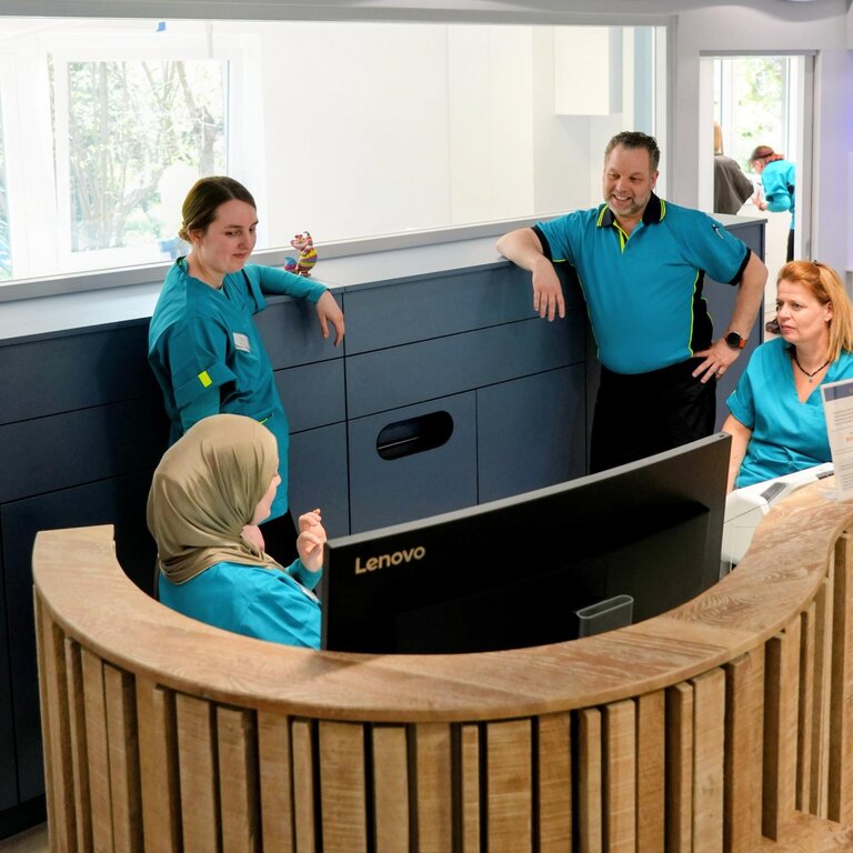Eine Gruppe von vier Personen in Arbeitskleidung steht an einem Empfangstresen. Zwei Personen blicken auf einen Computerbildschirm, während eine weitere Person auf sie schaut. Ein Fenster im Hintergrund lässt Tageslicht herein. Der Raum hat eine moderne, helle Gestaltung.