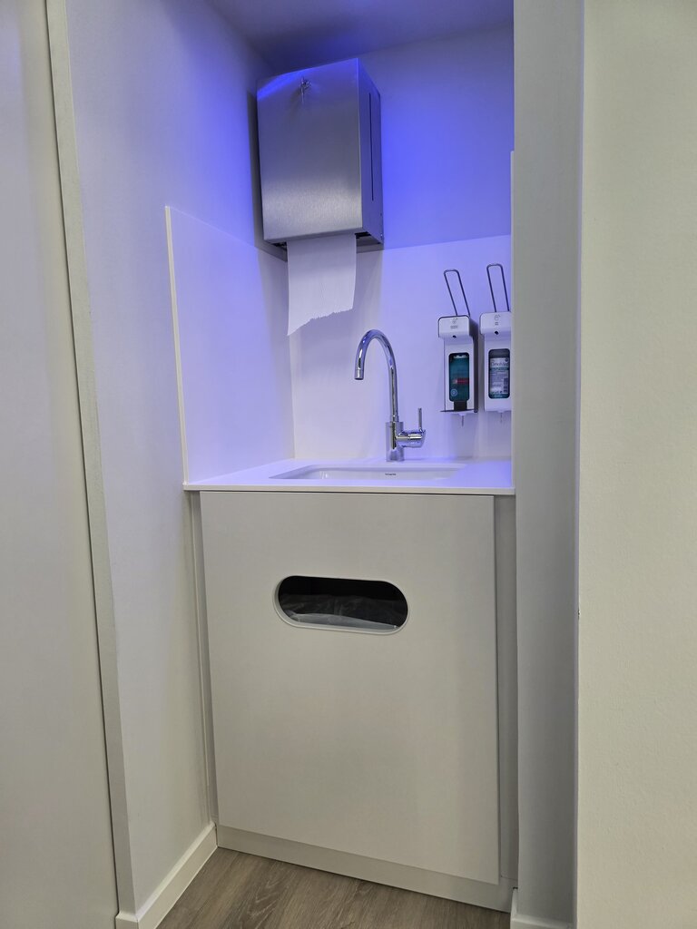 Ein minimalistischer Waschbereich mit einem Waschbecken, einem Wasserhahn und einem Seifenspender. Über dem Waschbecken befindet sich ein Papiertuchspender. Die Wand ist in einer hellen Farbe gehalten und es gibt eine indirekte Beleuchtung in Blau.