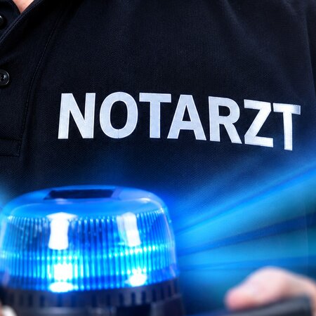 Ein Notarzt trägt ein schwarzes Hemd mit der Aufschrift "NOTARZT" und hält eine leuchtende Blaulichtlampe in der Hand. Die Umgebung ist unscharf, fokussiert auf die Person und das Blaulicht.