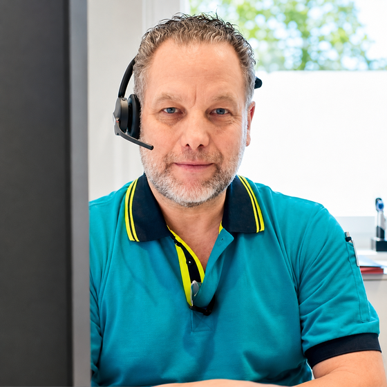 Ein Mann mit Headset sitzt an einem Arbeitsplatz. Er trägt ein turquoise T-Shirt mit schwarzen und gelben Akzenten. Im Hintergrund sind verschwommene Fenster und Büromaterialien zu sehen. Der Mann lächelt und schaut direkt in die Kamera.