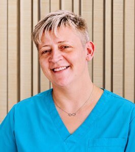 Eine lächelnde Person trägt ein hellblaues Oberteil. Der Hintergrund besteht aus hellen, vertikalen Linien. Die Person hat kurze, hellblonde Haare und sieht freundlich aus.