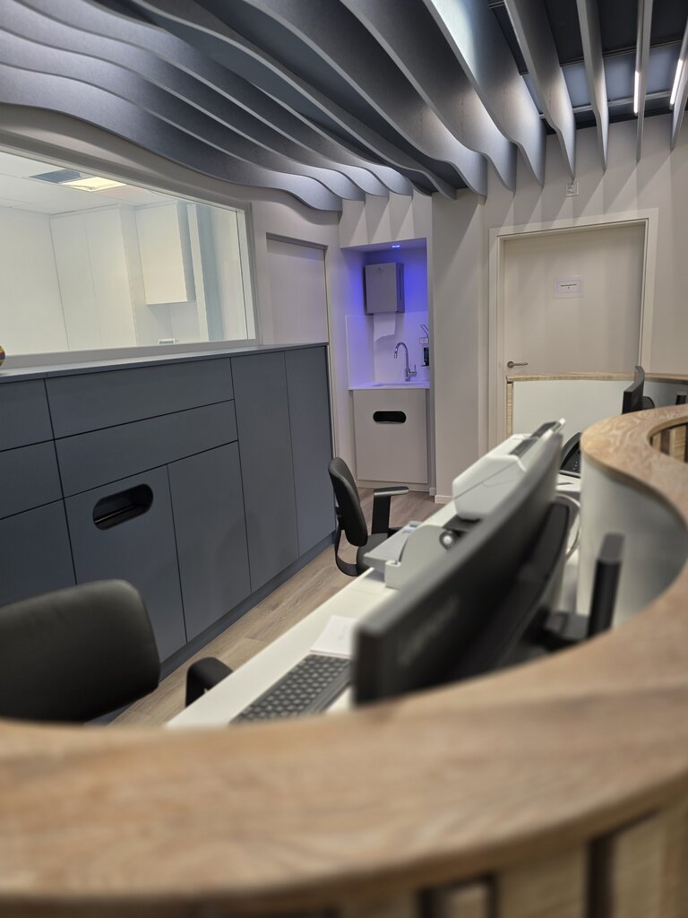 Moderne Büroumgebung mit einem Empfangstresen, mehreren Monitoren und ergonomischen Stühlen. An der Wand sind Regale und eine kleine, beleuchtete Küchenzeile zu sehen. Die Decke hat eine wellenförmige Gestaltung, die dem Raum ein modernes Ambiente verleiht.