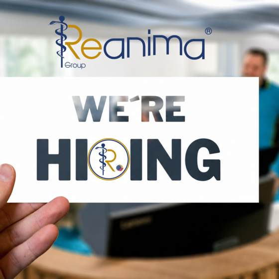 Ein Mensch hält ein Schild mit der Aufschrift "WE'RE HIRING" vor einem Empfangstresen, an dem Mitarbeiter in blauen Outfits stehen. Im Hintergrund ist der Schriftzug der Reanima Group zu sehen.