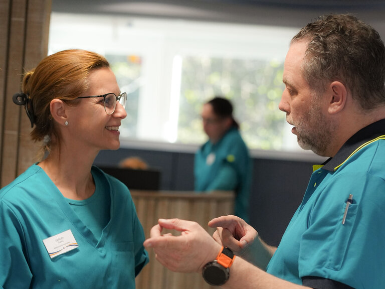 Zwei Pflegekräfte in blauen Uniformen unterhalten sich in einem hellen Raum. Die Frau mit Brille lächelt, während der Mann gestikuliert. Im Hintergrund ist eine weitere Person sichtbar. Die Atmosphäre wirkt freundlich und kollegial.