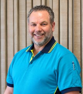 Ein lächelnder Mann mit kurzem, grauen Haar trägt ein hellblaues Polo-Shirt mit gelben und dunkelblauen Akzenten. Er steht vor einer Wand mit horizontalen, hellen Holzlatten und strahlt Selbstbewusstsein aus.