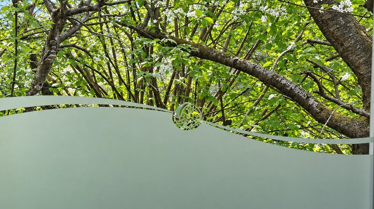 Eine grüne Glaswand zeigt im Hintergrund einen hell erblühten Baum mit vielen Ästen und frischem Laub. Die Lichtdurchlässigkeit des Glases erlaubt einen Blick auf die Natur, während die Wand eine sanfte, kurvige Form hat.