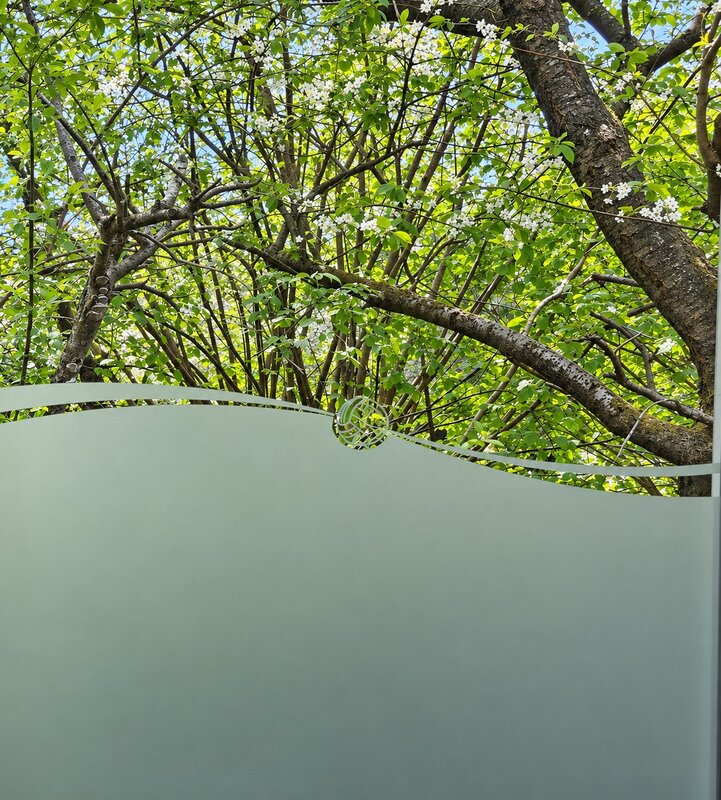 Eine grüne Glaswand zeigt im Hintergrund einen hell erblühten Baum mit vielen Ästen und frischem Laub. Die Lichtdurchlässigkeit des Glases erlaubt einen Blick auf die Natur, während die Wand eine sanfte, kurvige Form hat.